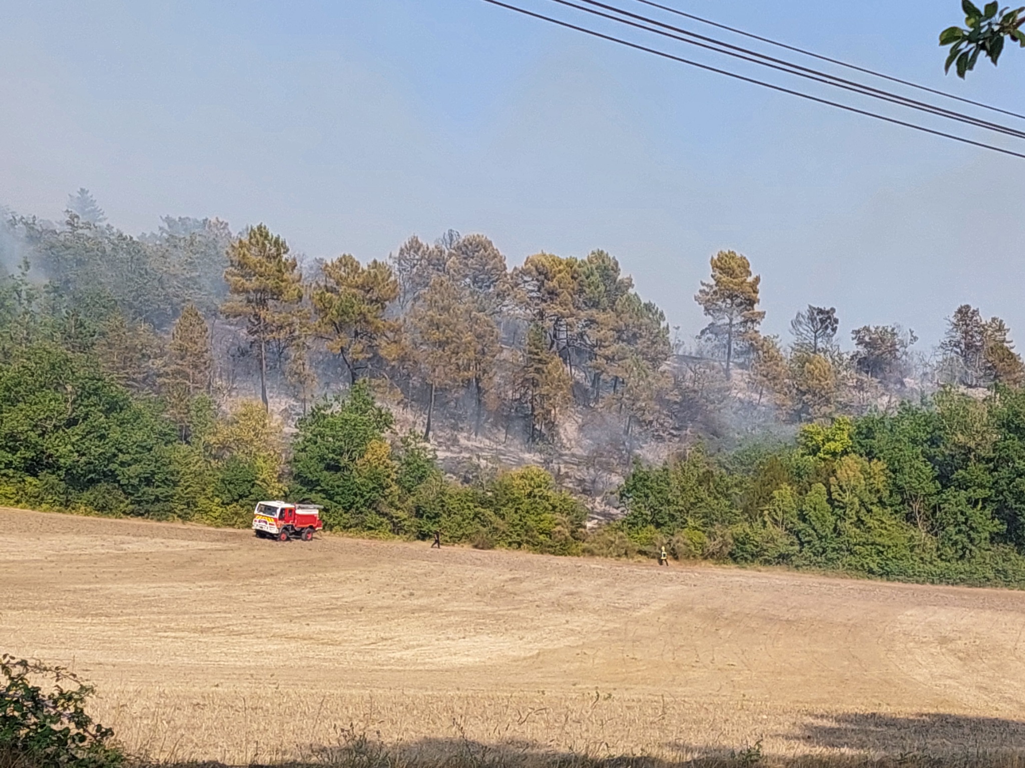 Wildfire near Laussou in Lot-et-Garonne, with a fire engine in a field below burned woodland.